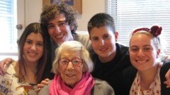 An elderly woman sits beside a table while her four teenage grandchildren crowd in close behind her