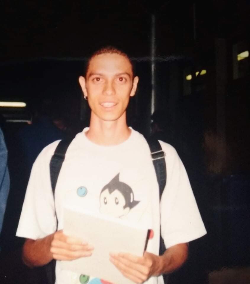 A grainy photo of a young Aboriginal man with a shaved head wearing an astroboy tshirt and backpack