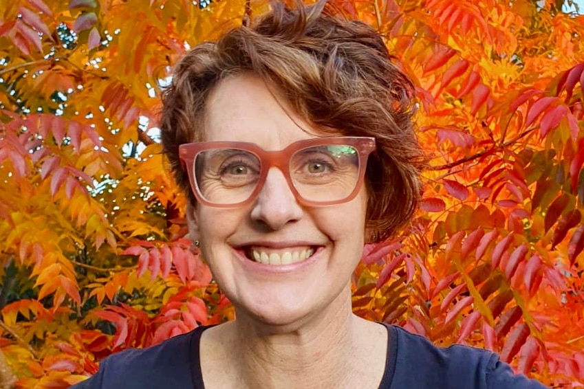 A profile of a woman with short, brunette hair and red reading glasses smiling widely.'