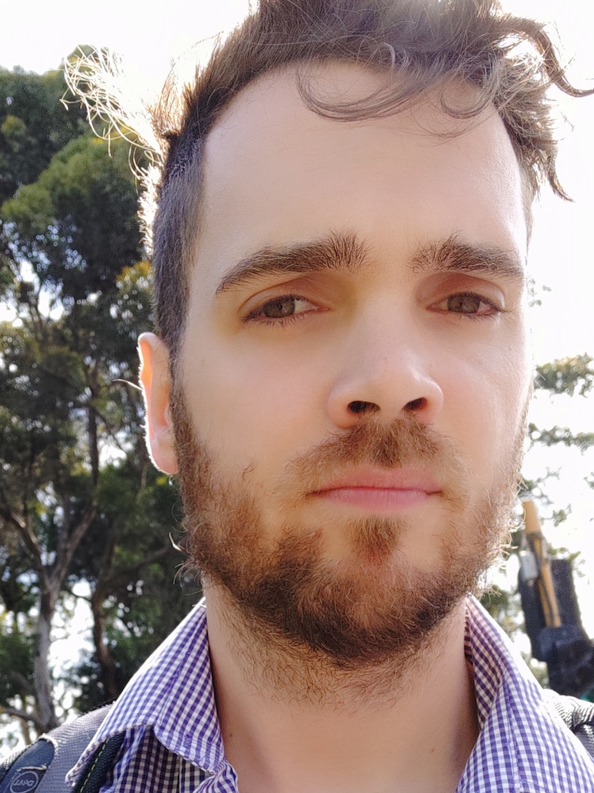 A young man with a short beard stands outside, looking at the camera.