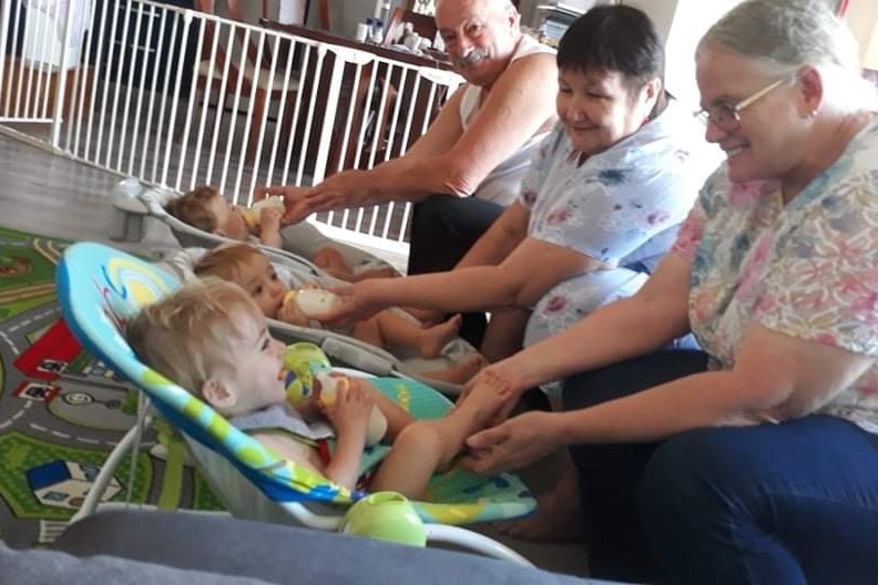 Three older people look down at three babies in bouncy chairs
