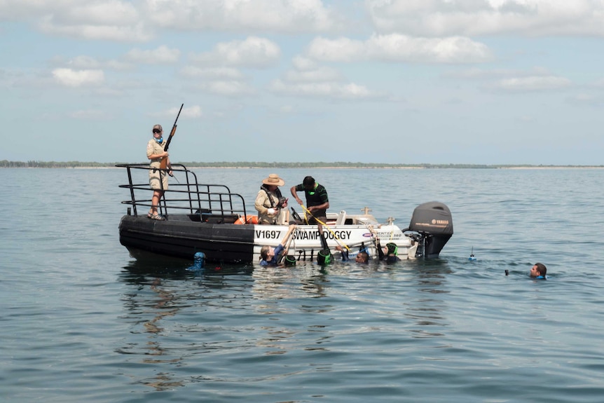 a woman stands with a gun in a boat while people in wetsuits measure a dugong on the side of the boat.