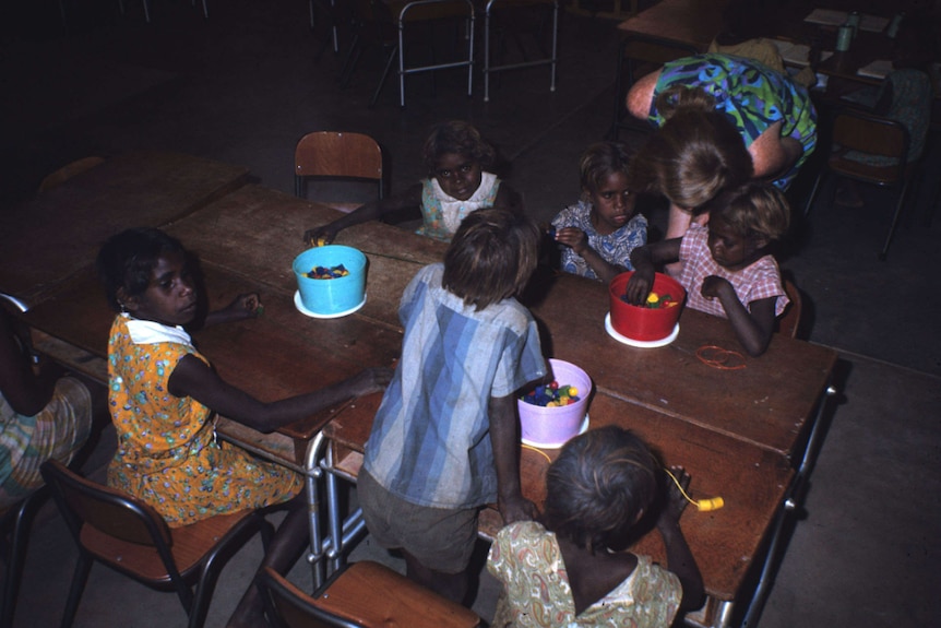 An old photo of children sitting at a table threading beads. A teacher leans over two kids