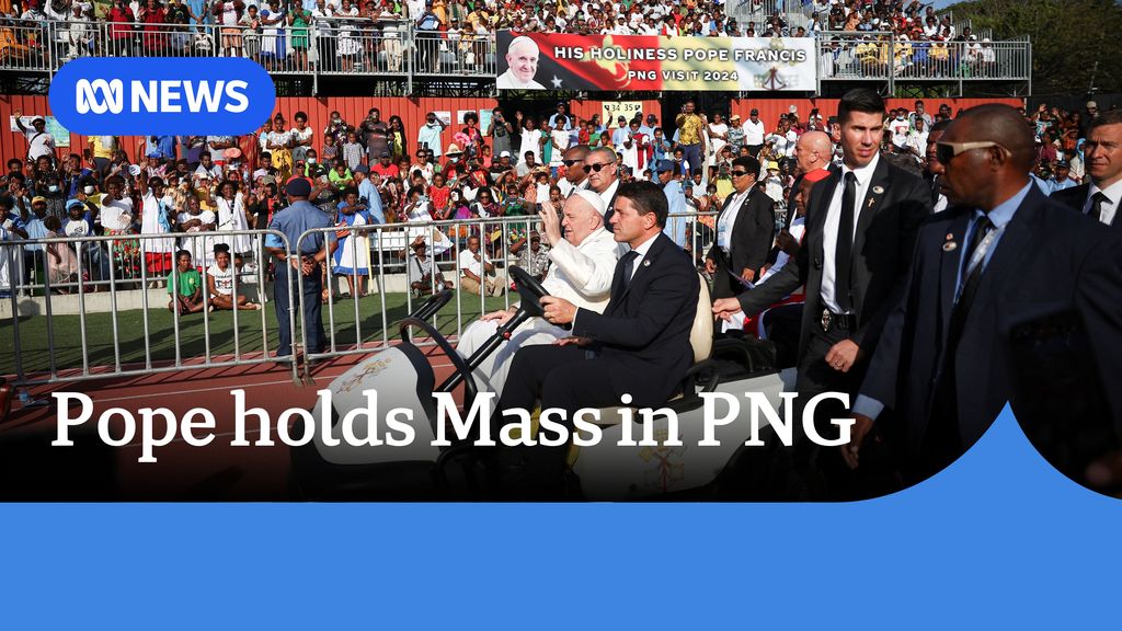 Pope holds Mass in PNG: The pope being driven in a white vehicle in a stadium with suited bodyguards escorting him.
