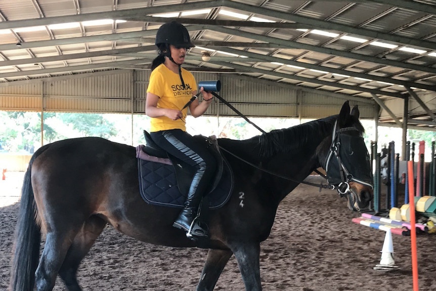 A girl in a yellow shirt rides a horse in a stable.