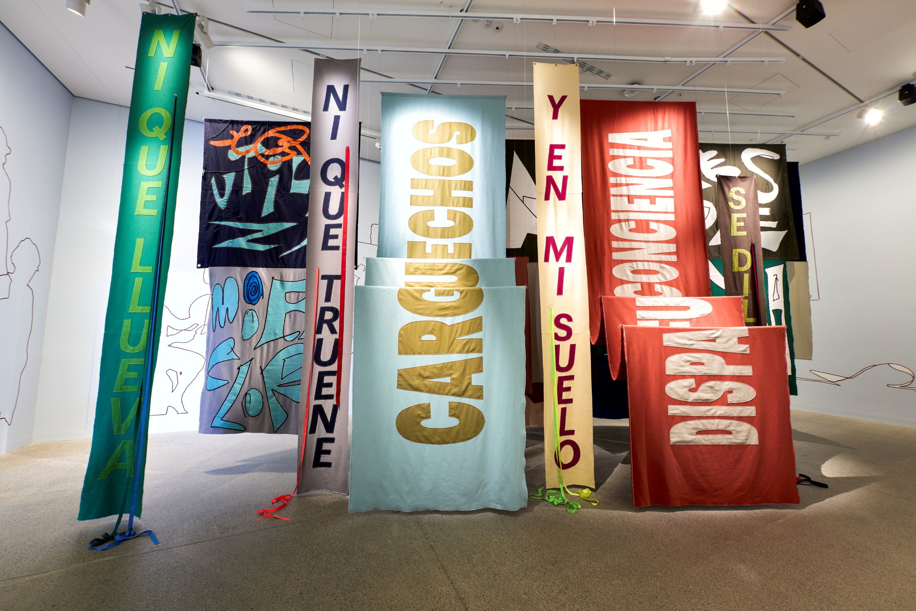 An artwork by Nadia Hernández; a series of colourful banners bearing slogans in Spanish, hung in a gallery.