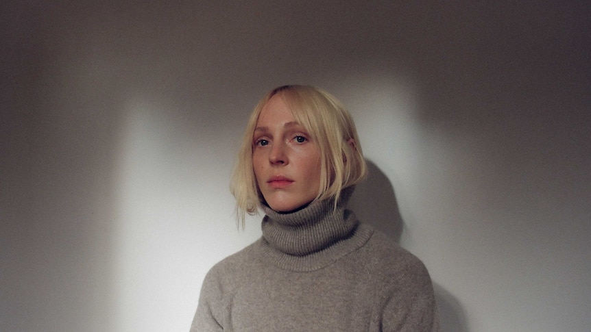 A blond woman has her hair tucked into a brown turtleneck jumper. She looks serious.