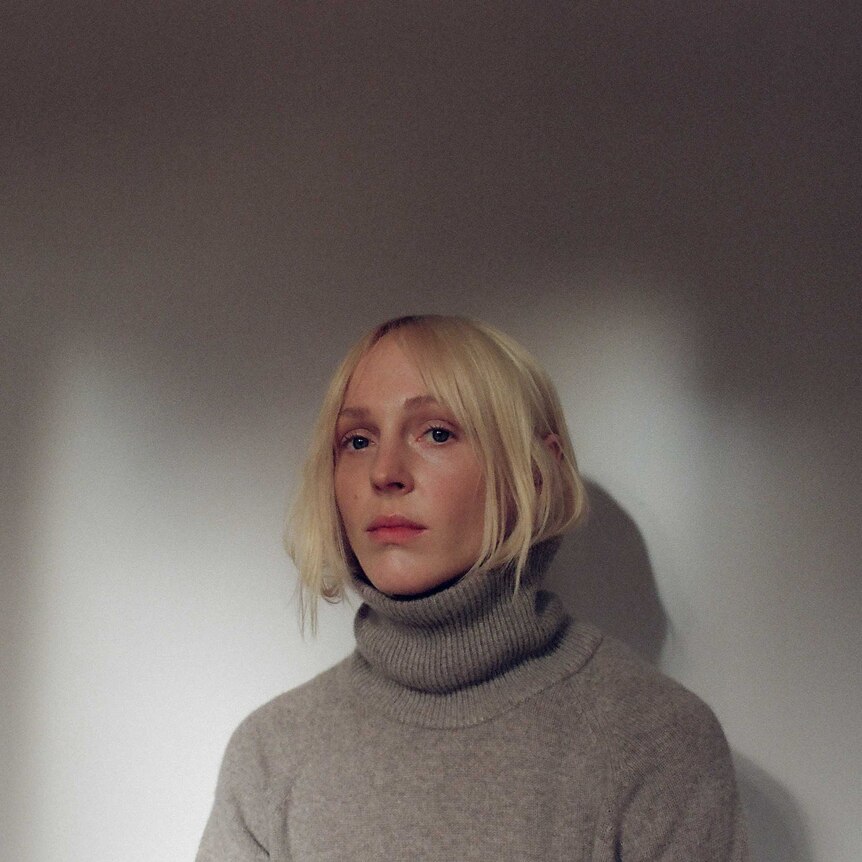 A blond woman has her hair tucked into a brown turtleneck jumper. She looks serious.