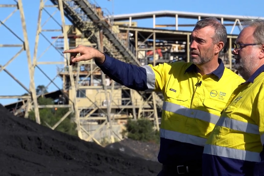 Two men in high vis standing in front of a coal pile with one of them pointing 