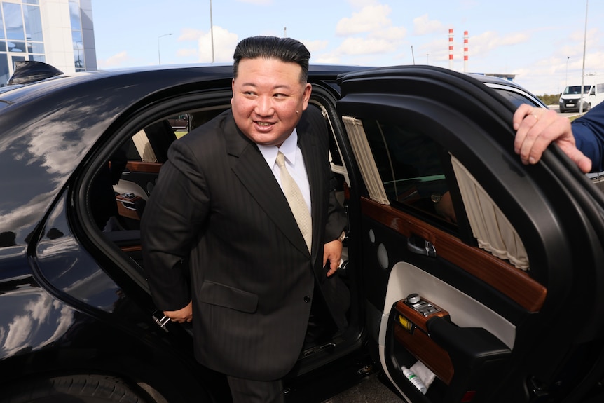 A man of Asian appearance wears a suit and steps out of a luxury car as its door is held open by someone else.