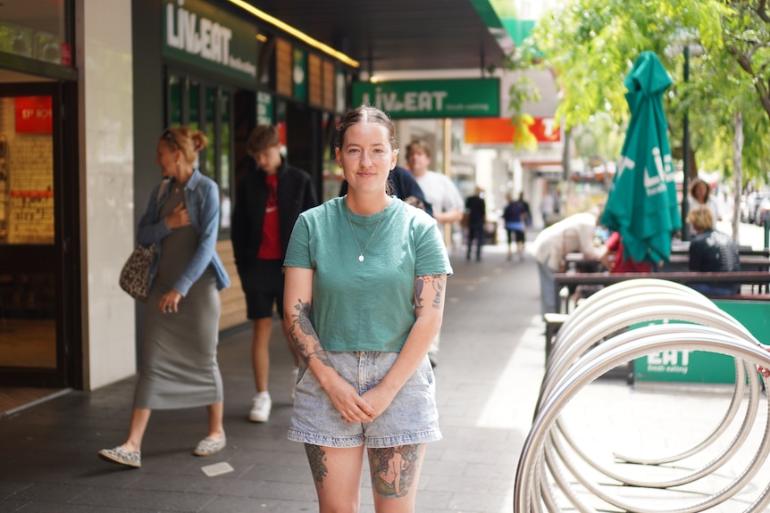 Una mujer joven con una camisa verde y tatuajes se encuentra en medio de un concurrido centro comercial, sonriendo a la cámara.