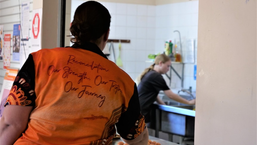 A woman in orange and black t-shirt has her back to camera, looks at a young girl in the kitchen.