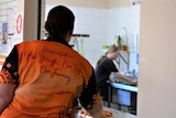 A woman in orange and black t-shirt has her back to camera, looks at a young girl in the kitchen.