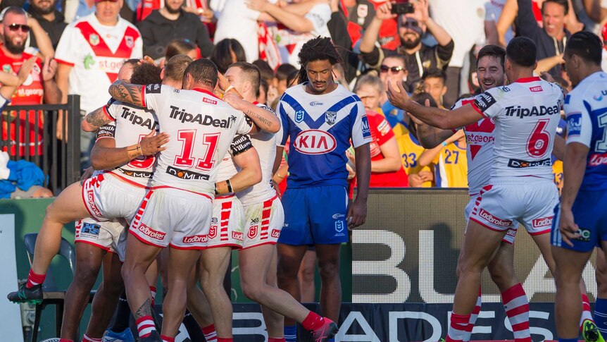 A rugby league team celebrates in a huddle after scoring a try as their opponents look down, dejected