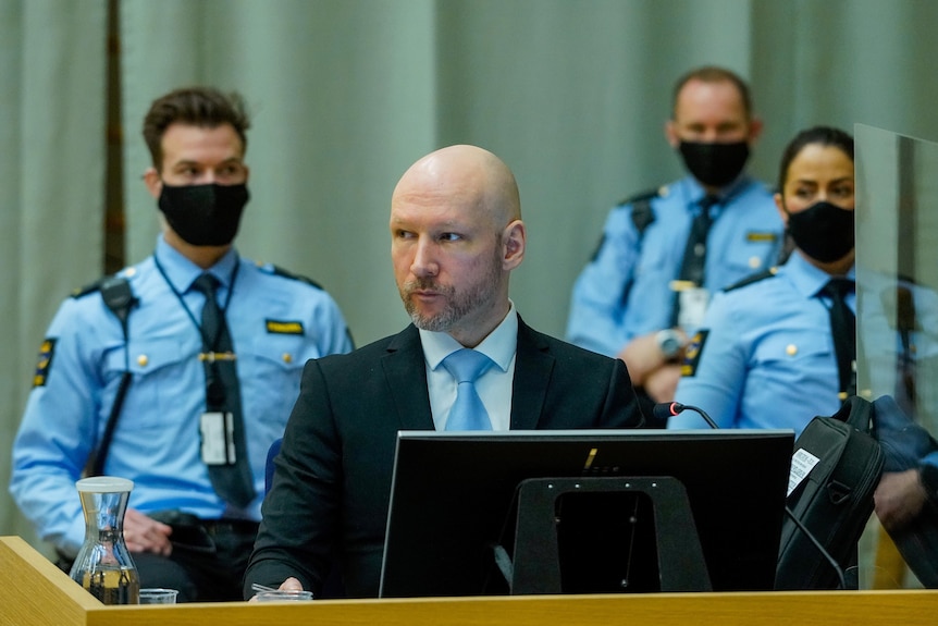 A bald man in a suit, with three masked security guards around him.