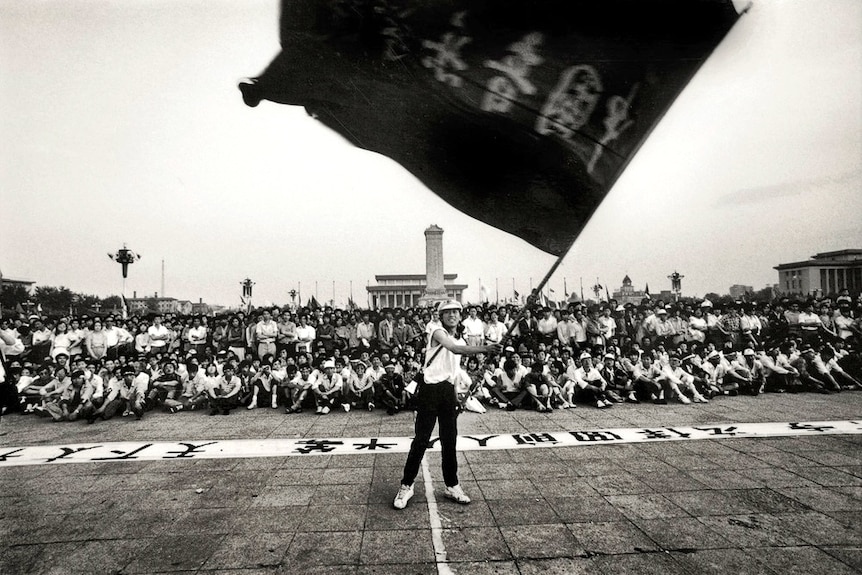 A black and white archival image shows a woman waving a large flag in front of thousands of people.