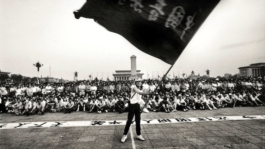 A black and white archival image shows a woman waving a large flag in front of thousands of people.