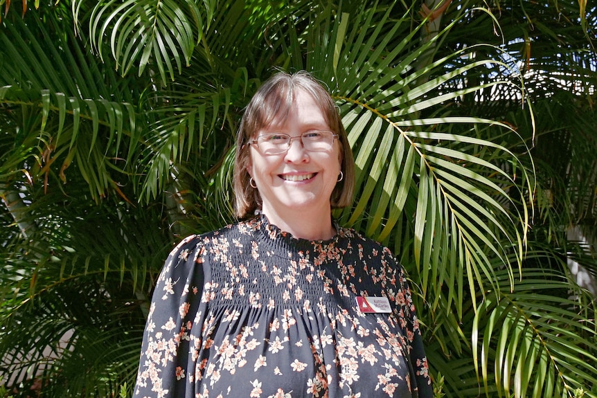 A woman wearing a black, floral shirt smiles, standing in front of green trees