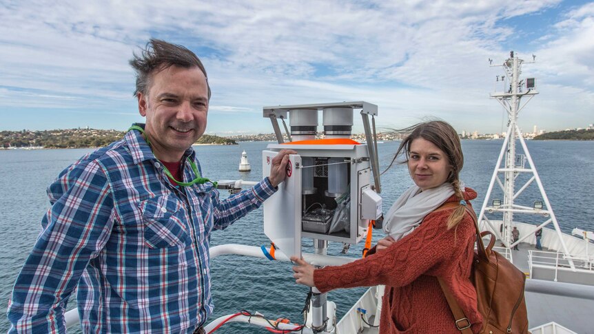 Two people stand with a piece of scientific equipment that looks like a metal fuse box. Sydney harbour is in the background.