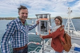 Two people stand with a piece of scientific equipment that looks like a metal fuse box. Sydney harbour is in the background.
