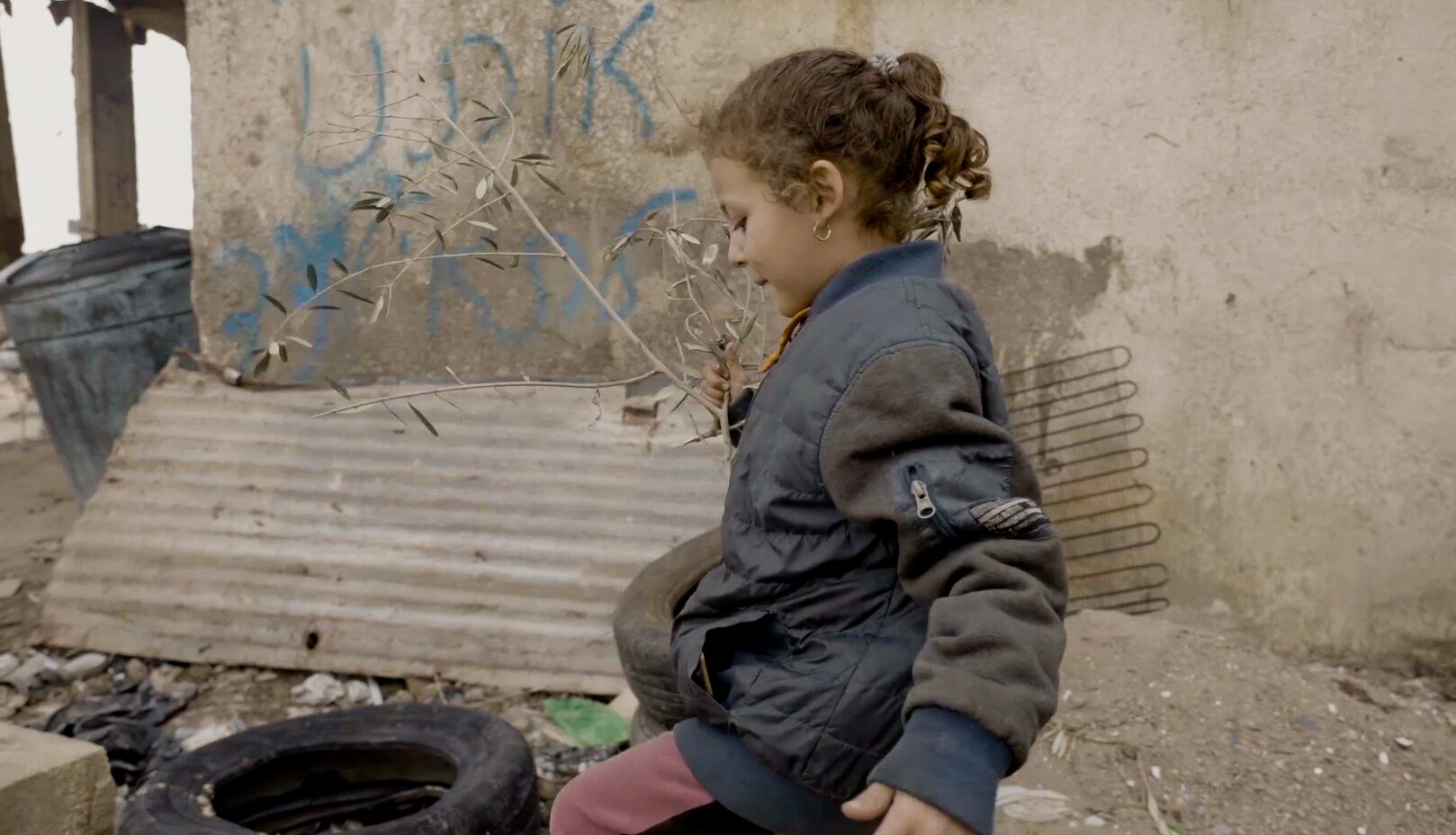 A girl wearing a coat carries a stick as she walks among the debris of her damaged home.