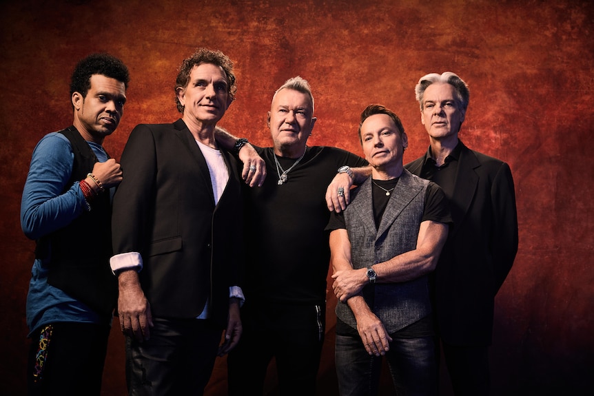 Cold Chisel band members stand in front of a deep orange backdrop. Five men standing in a row.