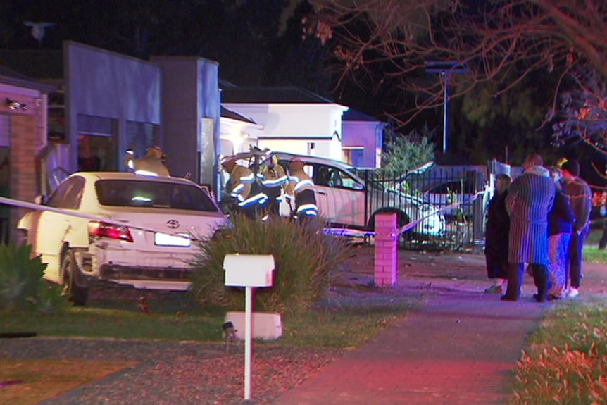 A damaged sedan in front of a house and a four-wheel drive and people watching at night