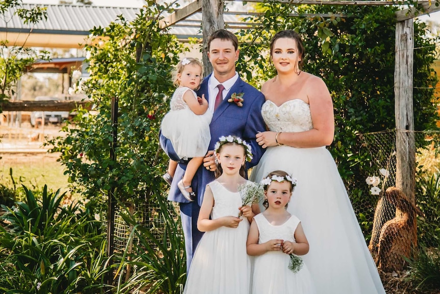 A bride and groom stand in front of greenery with their three young daughters, smiling.