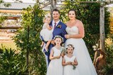 A bride and groom stand in front of greenery with their three young daughters, smiling.