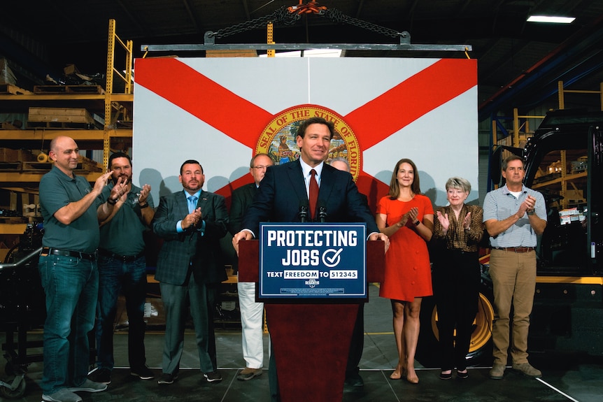 Ron DeSantis in a blue suit and red tie stands in front of the Florida flag, behind a podium that says "protecting jobs"
