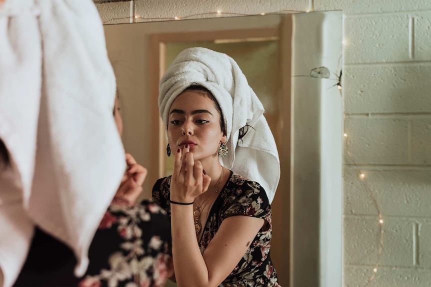 Young woman fixing her make-up in front of a mirror with a towel on her hair to depict how often to wash everyday items.