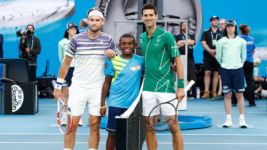 A 10-year-old boy stands at the tennis net with Dominic Thiem and Novak Djokovic at the Australian Open
