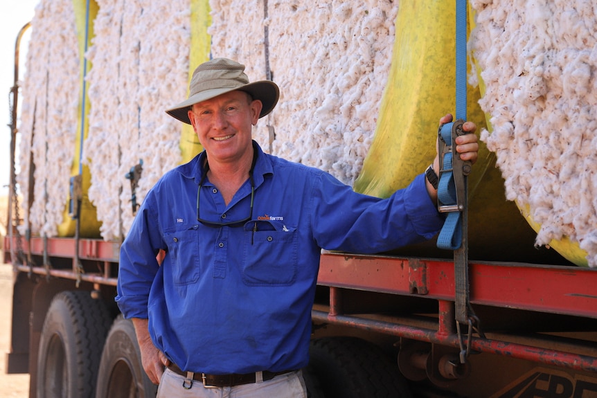 A man in a long-sleeved blue shirt, beige trousers and a broad-brimmed hat stands next to a truck with cotton bales.