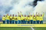 Australia's women's rugby sevens team stand on podium waving with their gold medals as firework smoke drifts behind them. 