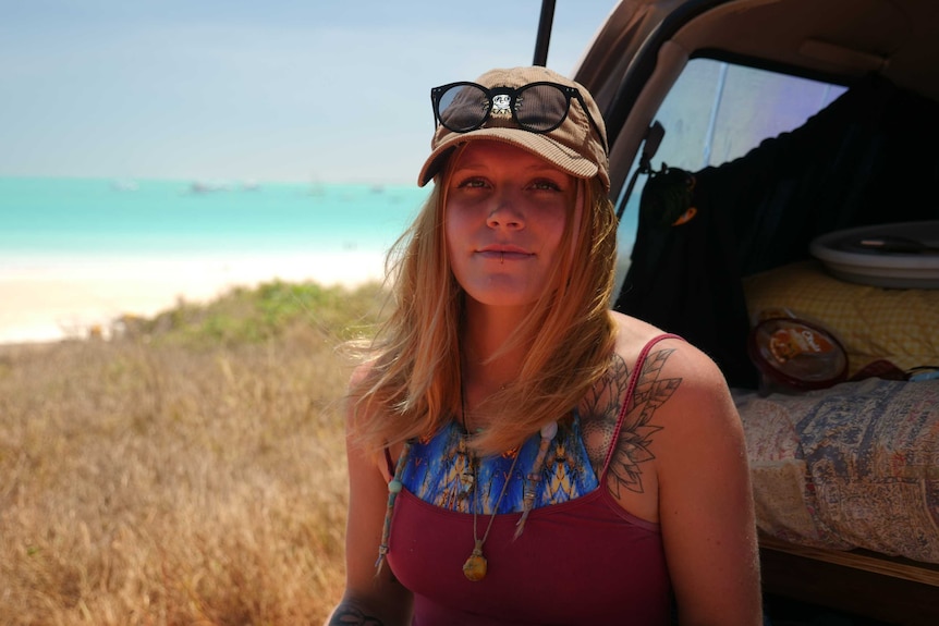 A young woman with  a hat and sunglasses on sits on the boot of her car in Broome with luggage packed