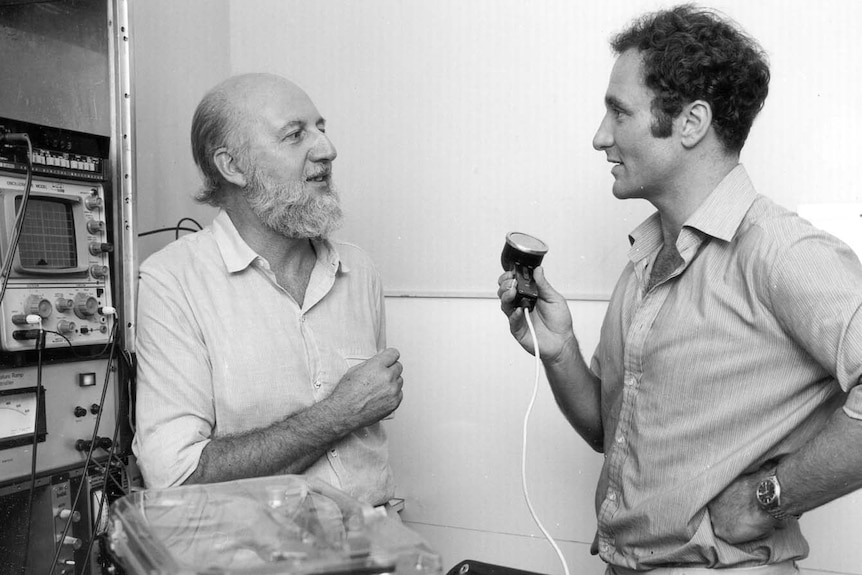 Black and white photo of man holding old fashioned radio microphone interviewing another man next to audio equipment.