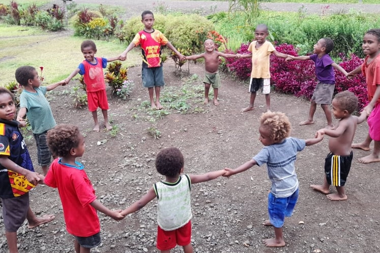 Children in a rustic setting stand in a circle, holding hands.