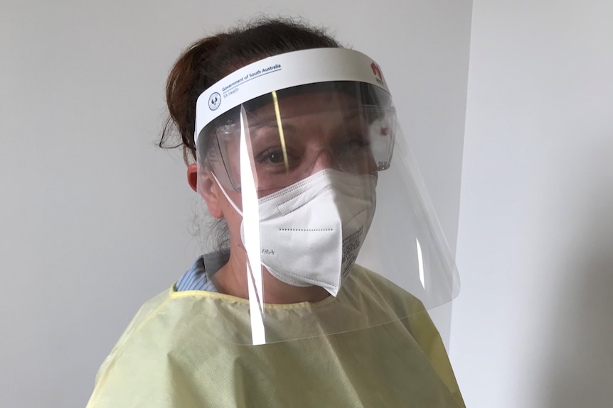 A female nurse with hair in a bun wears a mask, face shield and yellow gown.
