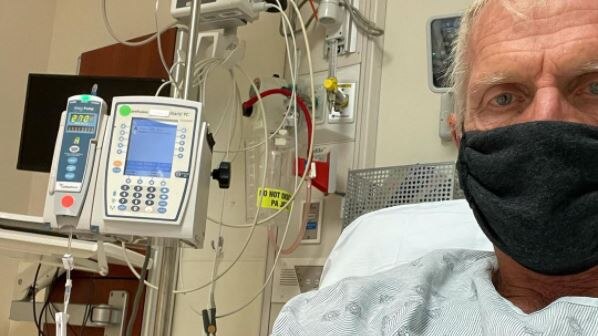 Greg Norman sits in a hospital bed with a drip in his arm and a black face mask on after testing positive for COVID-19.