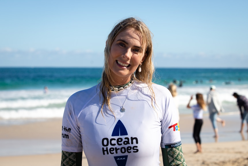 A blonde woman in a white rashie and green wetsuit smiles at the camera.