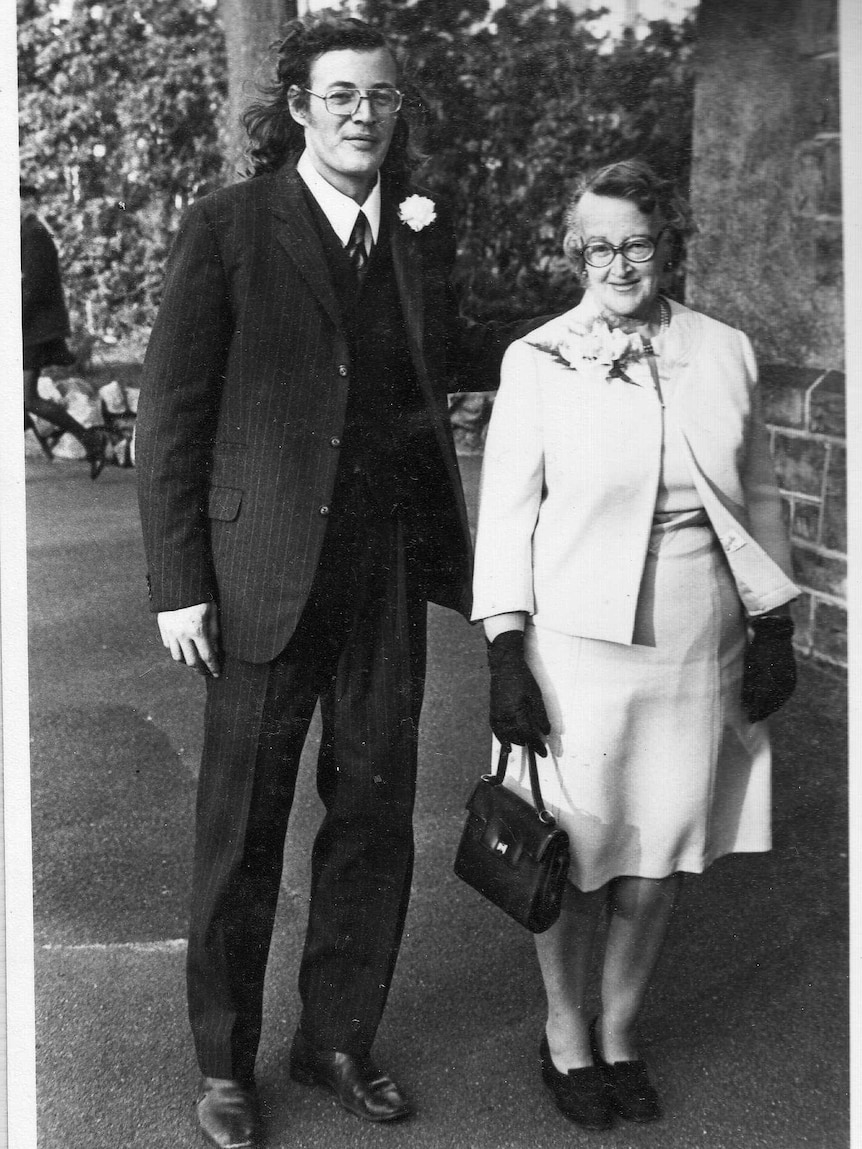 A mother and adult son in formal attire. Son wears dark suit and tie with flower, mother wears white with white flower