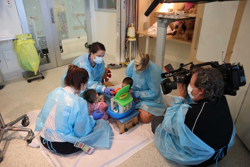 Cameraman in blue gown and wearing face mask films hospital staff similarly dressed playing with twins and toys.