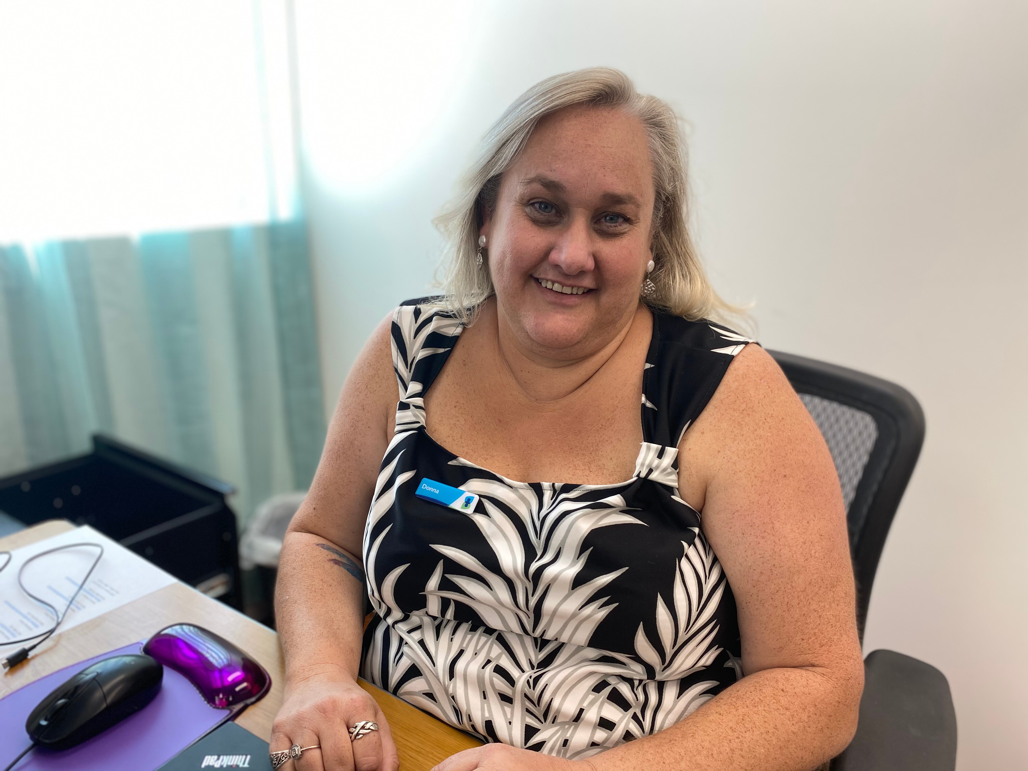A woman, wearing a badge, sits smiling at her desk
