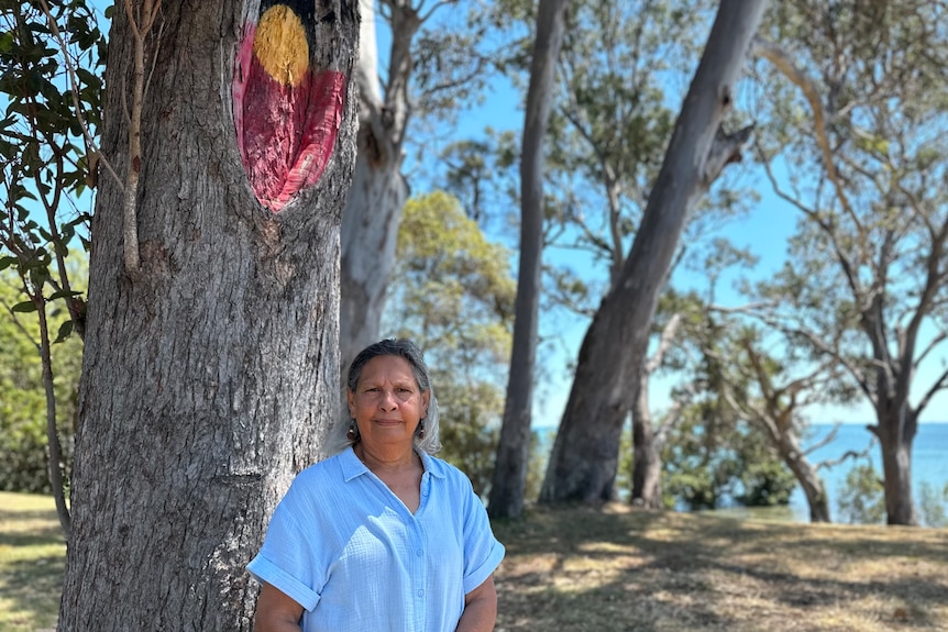 A woman in a light blue shirt stands in front of a tree with an Indigenous flag painted into a knot
