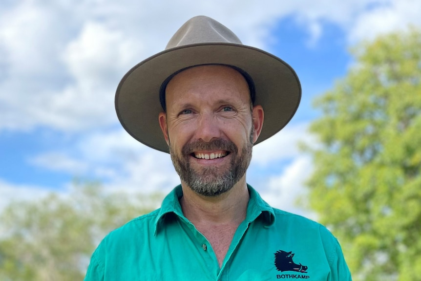 Photo de tête d'un homme avec une chemise verte et un chapeau à large bord marron et une barbe noire souriant.