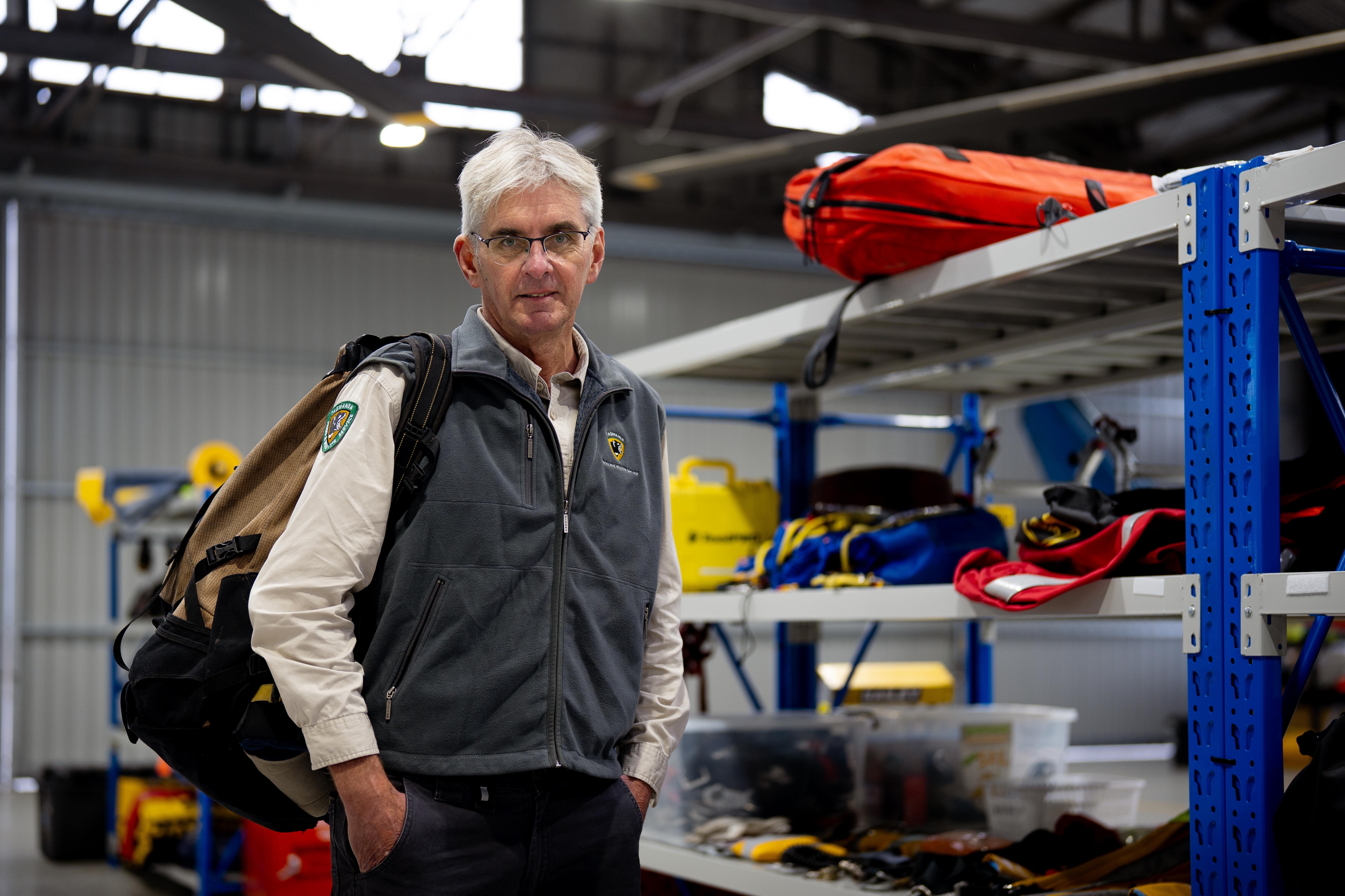 Brendan Moodie, wearing a cream shirt with a Tas Parks patch and a backpack, stands beside a shelf of rescue gear.