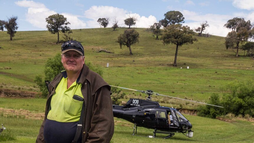 A farmer standing in drizabone in front of an ABC tv helicopter which has landed in his paddock