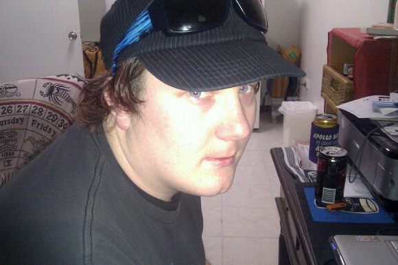 A young man wearing a dark cap and T-shirt sits at a computer desk.