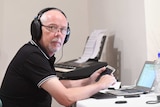 Dan McGarry looks at the camera with headphones on while holding a phone, in front of a laptop.