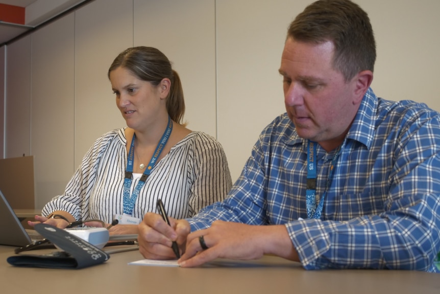A man and a woman, both paramedics, write health assessments for patients. One writes on paper, the other types into a laptop